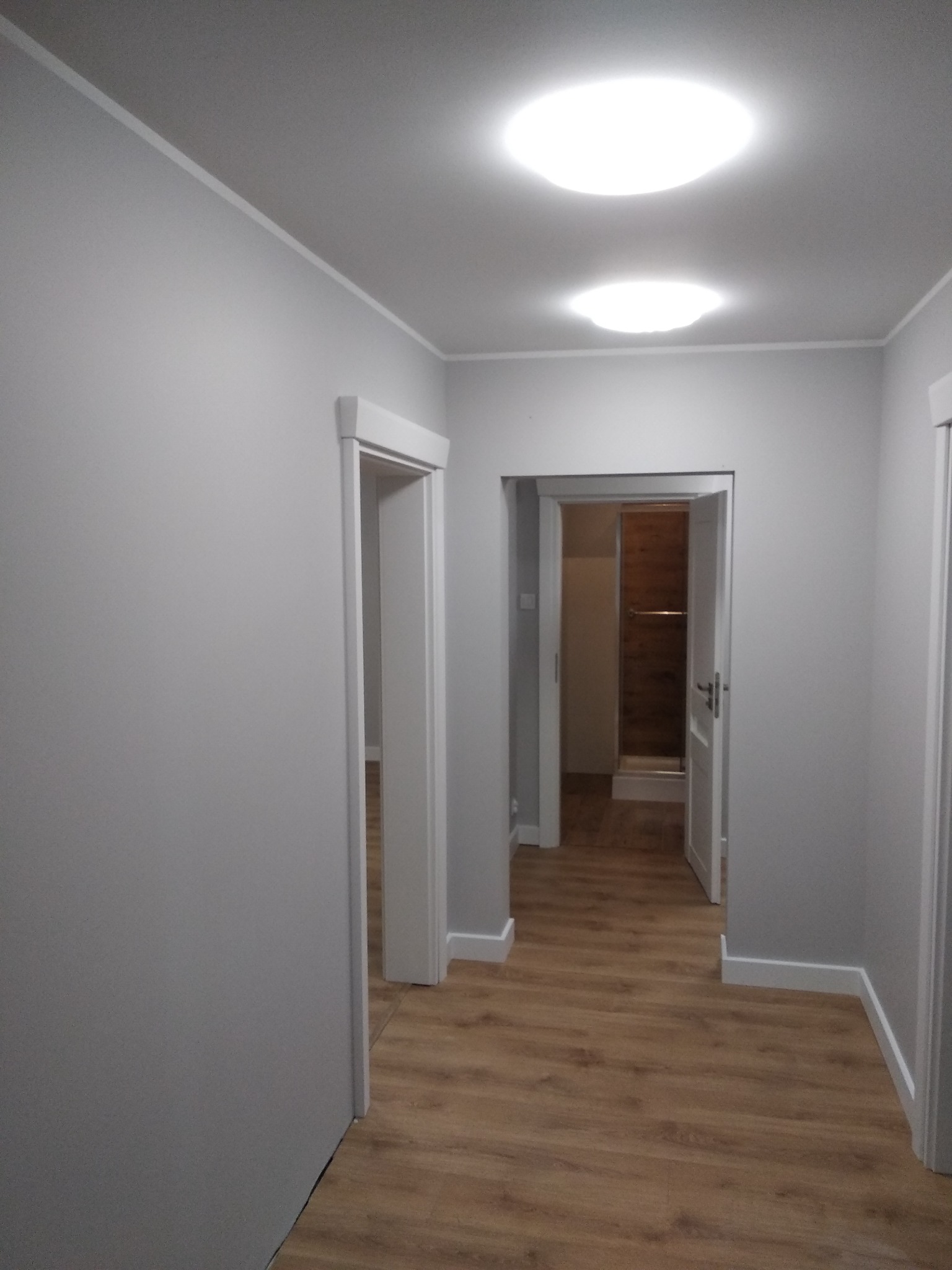 Szary korytarz z drewnopodobną podłogą, białymi listwami przypodłogowymi i trzema parami drzwi, z których jedne prowadzą do wykończonej łazienki z kabiną prysznicową.