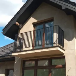 Nowoczesny balkon z czarną, prostą balustradą metalową, widok z dołu na tle błękitnego nieba z chmurami.