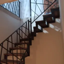 Nowoczesne schody metalowe z drewnianymi stopniami i geometryczną balustradą, widok z dołu na tle białej ściany i fragmentu ściany z cegły.