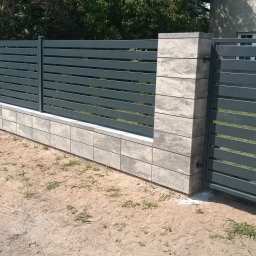 Z.P. KONSTAL - Nowoczesne ogrodzenie z poziomych paneli w kolorze grafitowym, osadzone na betonowej podmurówce z bloków o fakturze kamienia, z bramką wejściową, na tle piaszczystego podłoża i domu.