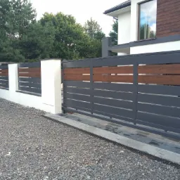 Nowoczesna, przesuwna brama wjazdowa z szarych, metalowych profili z poziomymi wstawkami z drewna, osadzona między białymi, murowanymi słupkami na tle domu i zieleni.