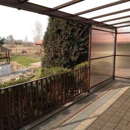 Z.P. KONSTAL - Zadaszony taras z brązową metalową balustradą i ścianami z przezroczystego tworzywa sztucznego, widok na ogród z altaną.