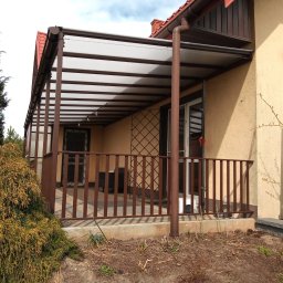 Z.P. KONSTAL - Zadaszony taras z brązowymi metalowymi balustradami i przezroczystym zadaszeniem, widok z zewnątrz na tle iglastego drzewa.