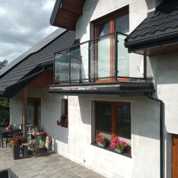 Z.P. KONSTAL - Nowoczesny dom z balkonem zabezpieczonym szklaną balustradą z czarnymi elementami mocującymi, widok z boku na elewację z oknami i kwiatami w doniczkach.