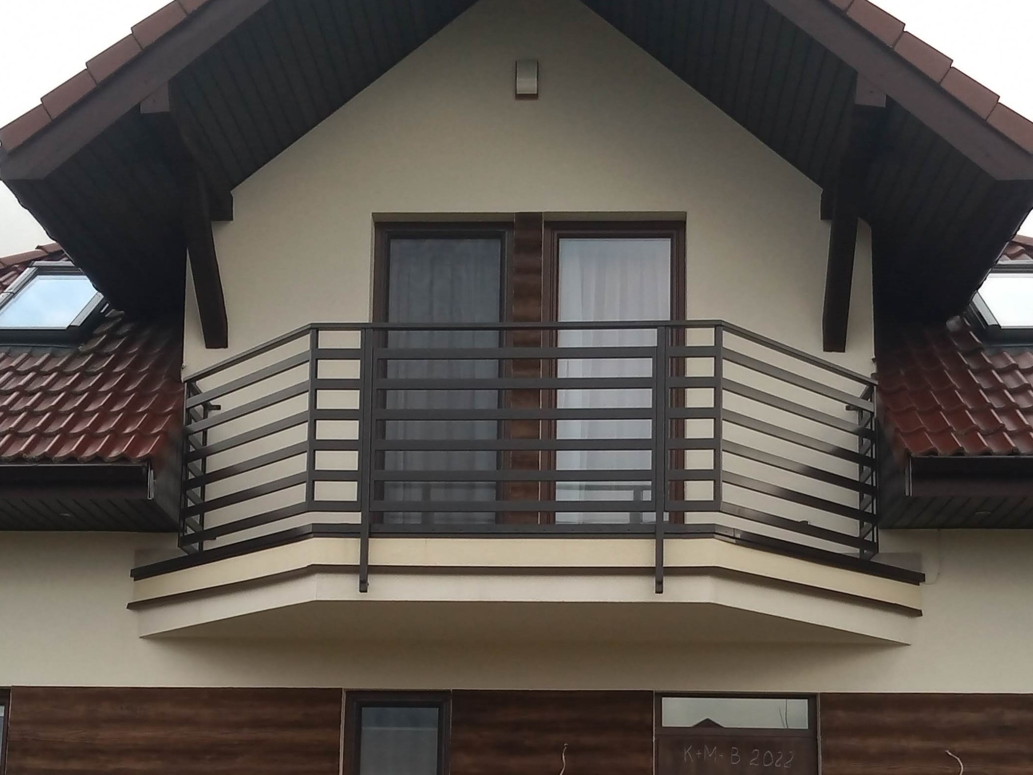 Metalowa balustrada balkonowa z poziomymi szczeblami, zamontowana na balkonie domu jednorodzinnego z brązową dachówką i oknami dachowymi.
