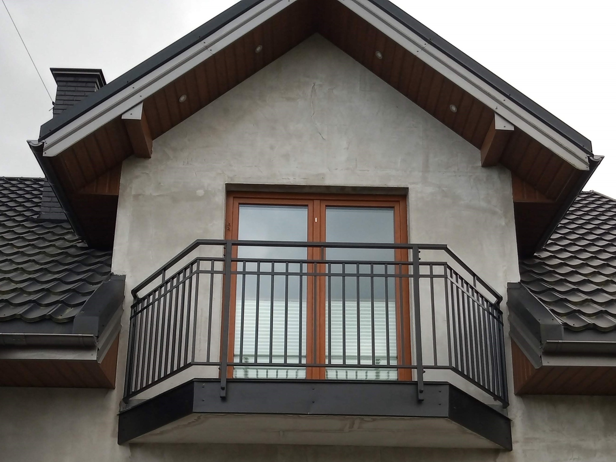 Czarna, metalowa balustrada balkonowa z prostymi szczeblami, zamontowana przed dwuskrzydłowymi drzwiami balkonowymi w domu z szarą elewacją i ciemną dachówką.