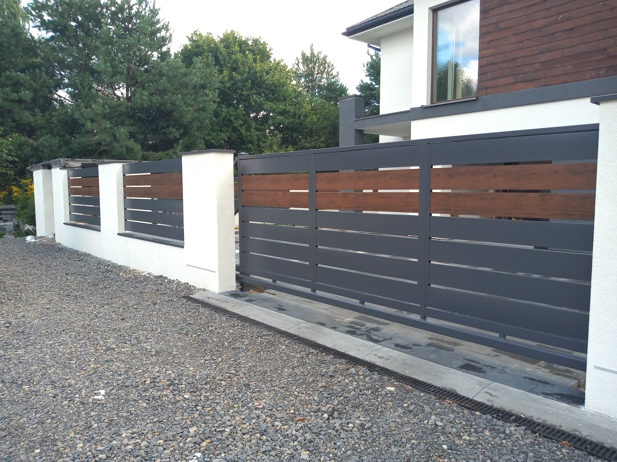 Nowoczesna, przesuwna brama wjazdowa z szarych, metalowych profili z poziomymi wstawkami z drewna, osadzona między białymi, murowanymi słupkami na tle domu i zieleni.
