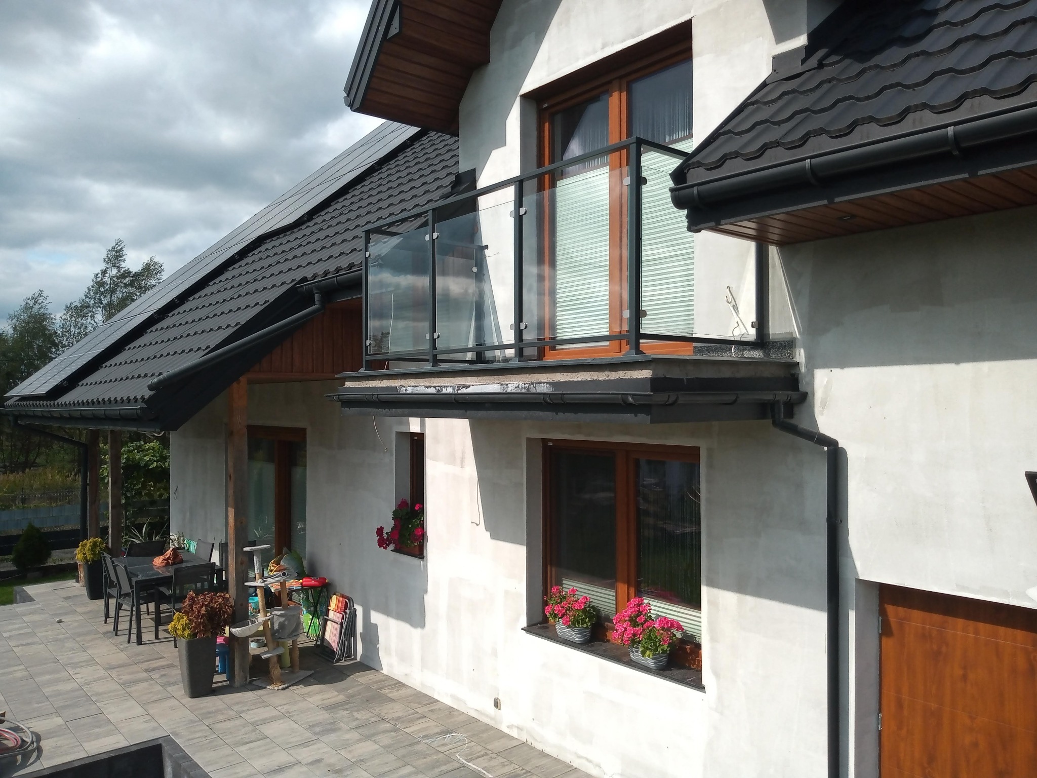 Nowoczesny dom z balkonem zabezpieczonym szklaną balustradą z czarnymi elementami mocującymi, widok z boku na elewację z oknami i kwiatami w doniczkach.