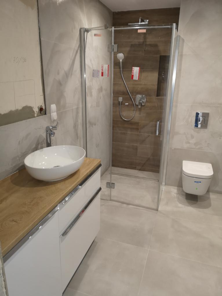 Nowoczesna łazienka z kabiną prysznicową, umywalką nablatową na szafce z drewnianym blatem i podwieszaną toaletą. Część ściany z widocznymi śladami prac wykończeniowych.