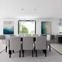 Jasna jadalnia z czarnym stołem, szarymi krzesłami, abstrakcyjnymi obrazami na ścianach, oświetleniem punktowym i widokiem na zieleń za oknem.