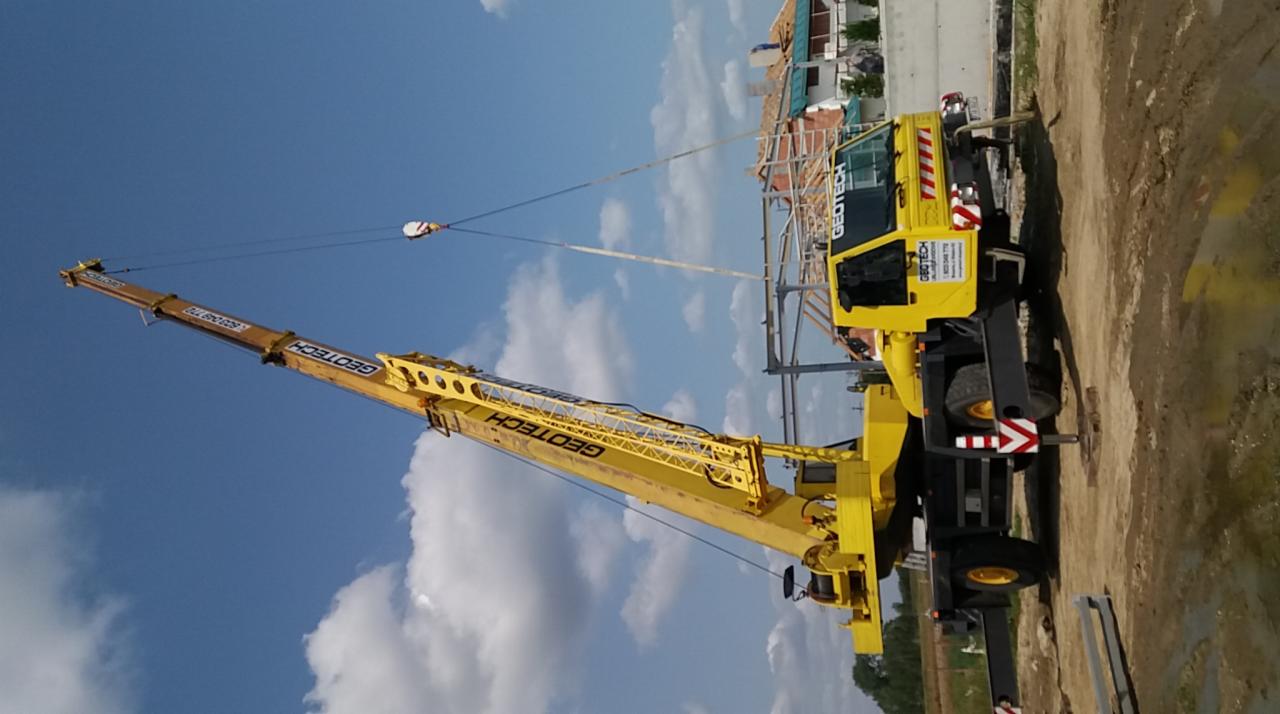 Żółty dźwig samojezdny marki Geotech z wysięgnikiem teleskopowym, ustawiony na placu budowy. W tle widoczne elementy konstrukcji budynku i błękitne niebo z chmurami.