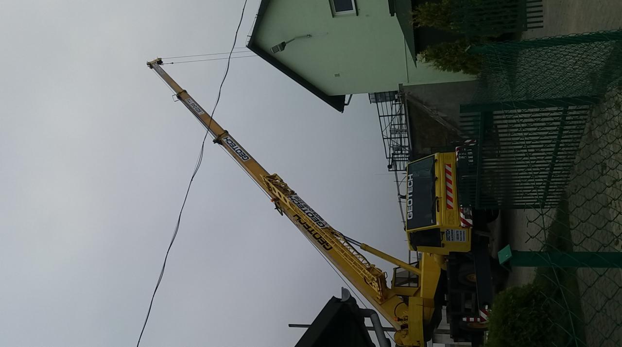 Żółty podnośnik koszowy marki Geotech z wysuniętym ramieniem na tle szarego nieba i budynku. Ujęcie z dołu.