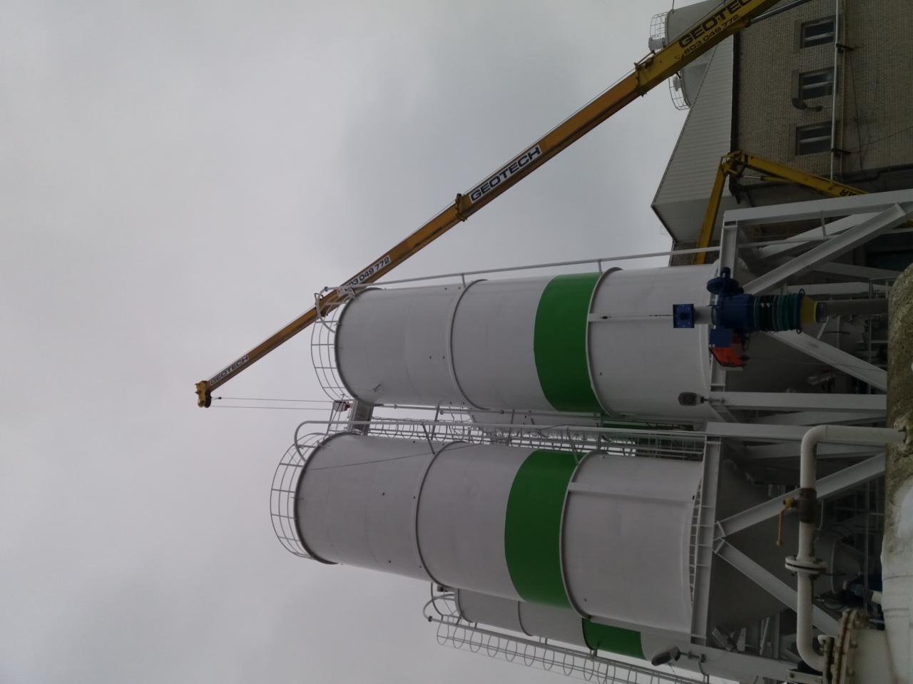 Dwa białe zbiorniki z zielonymi pasami, umieszczone na stalowej konstrukcji, widziane z dołu w pochmurny dzień, z dźwigiem GEOTECH w tle i fragmentem ceglanego budynku.