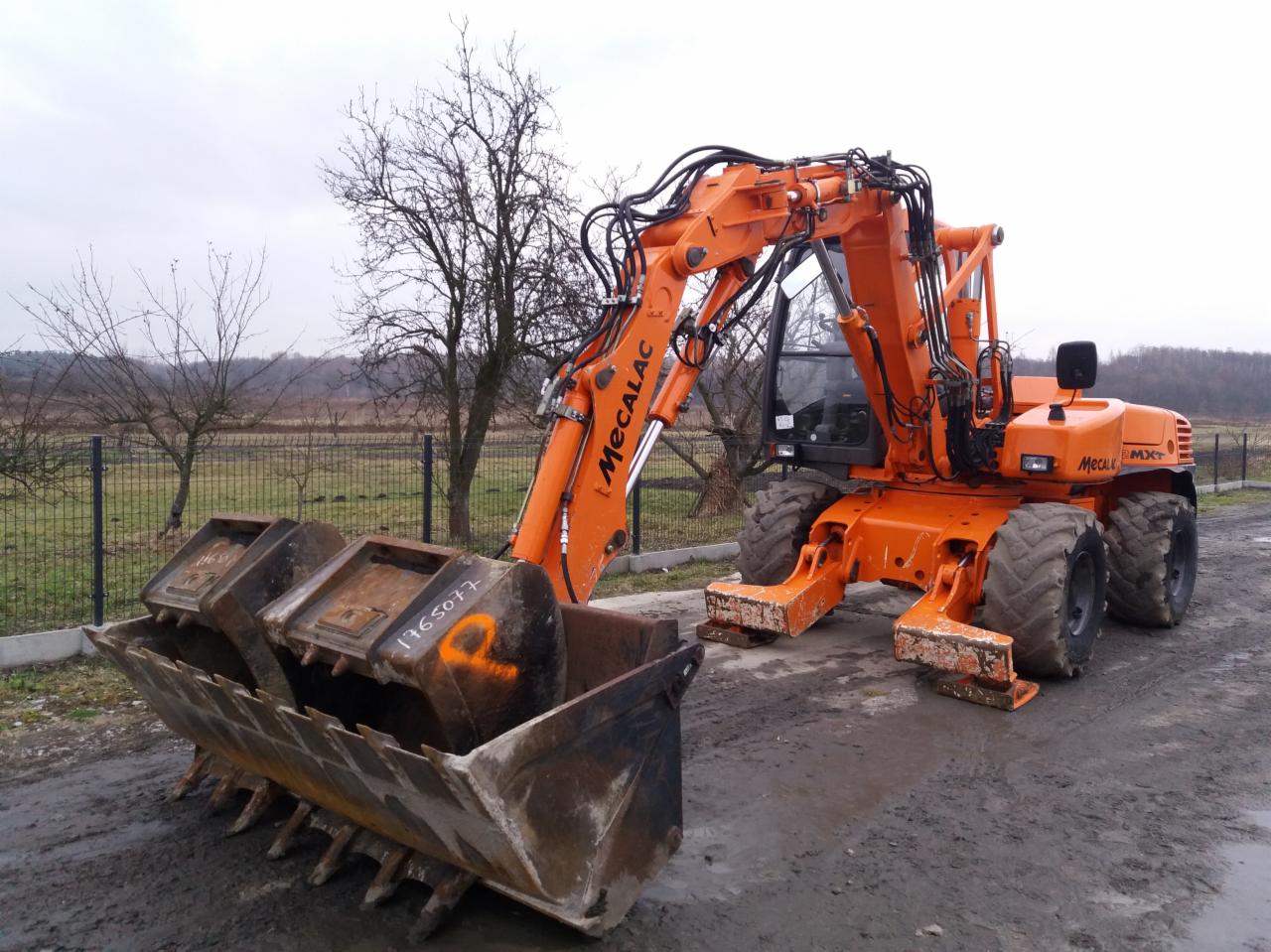 Pomarańczowa koparka kołowa Mecalac z trzema łyżkami na błotnistym placu, widoczne drzewa w tle i pochmurne niebo.