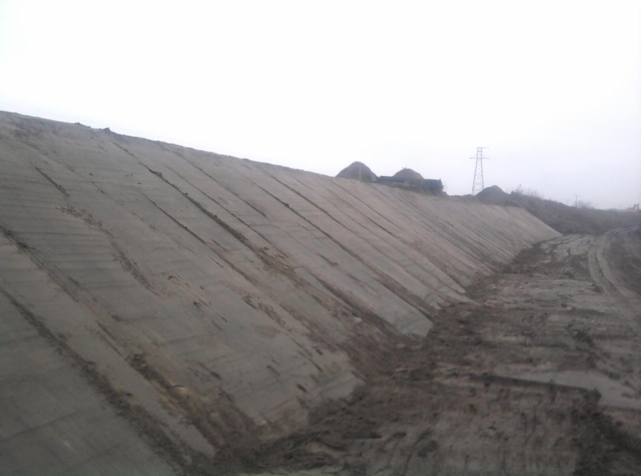 Uformowany, betonowy nasyp ziemny z widocznymi śladami po maszynach, w tle słup wysokiego napięcia i hałdy ziemi, pochmurny dzień.