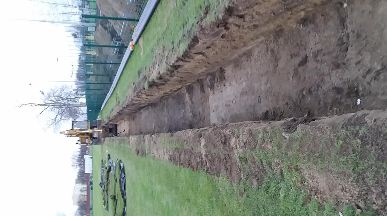 Długi wykop w ziemi na trawiastym terenie, z widocznym żółtym minikoparką pracującą w oddali, czarne folie rozłożone na trawie obok wykopu, ogrodzenie z siatki w tle.