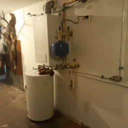 Biały piec gazowy dwufunkcyjny z niebieskim zbiornikiem wyrównawczym, widoczne rury miedziane i plastikowe, instalacja elektryczna, tło stanowi ściana z widocznymi śladami po instalacjach,...