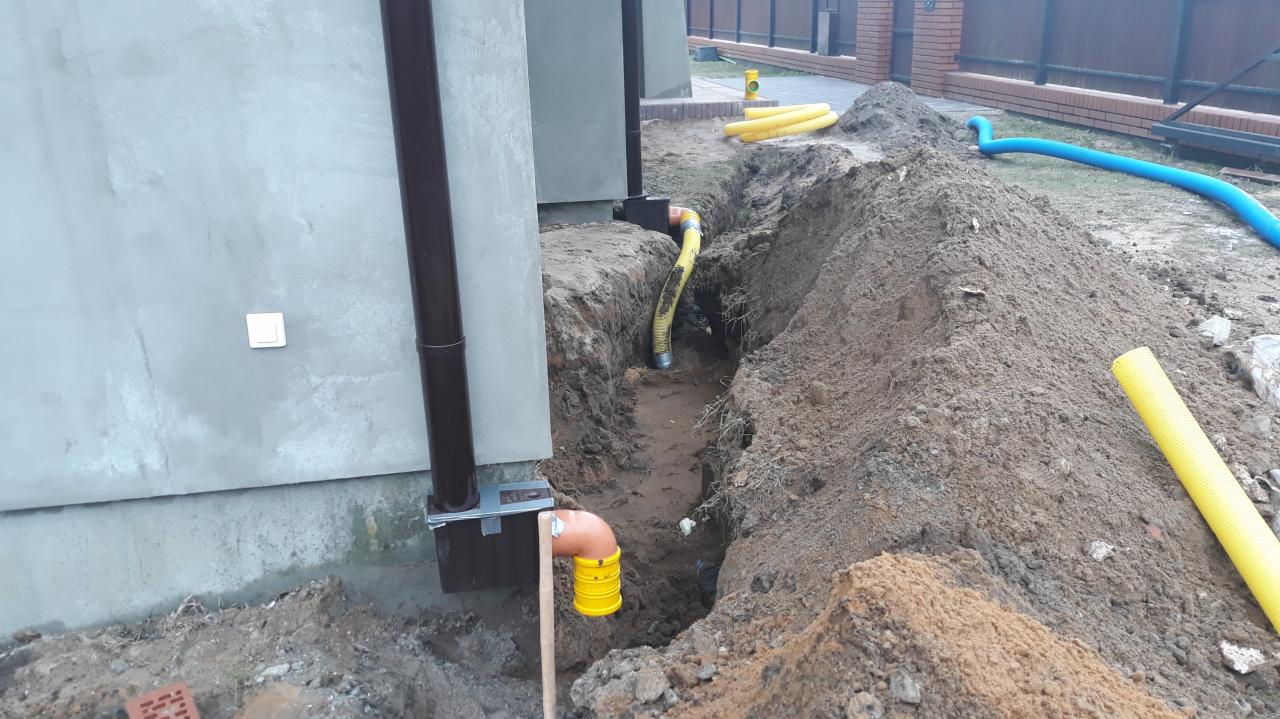 Wykop z widocznymi żółtymi rurami karbowanymi i pomarańczową kształtką kanalizacyjną przy fundamencie budynku, instalacja podziemna w trakcie realizacji.