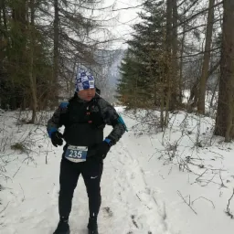 Biegacz z numerem 235 w zimowym lesie. Mężczyzna w sportowym stroju i czapce w niebiesko-białą kratę stoi na śnieżnej ścieżce, otoczony drzewami.