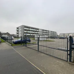 Otwarta brama wjazdowa z paneli ogrodzeniowych na posesję z blokiem mieszkalnym w tle, widoczny automat do bramy, szara kostka brukowa i zaparkowane samochody.