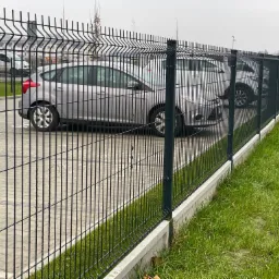 Ciemnozielone ogrodzenie z paneli na betonowej podmurówce, oddzielające parking z samochodami od trawnika w pochmurny dzień.