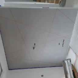 Świeżo położone gładzie na suficie, wyznaczone linie pod dekoracyjne malowanie, widoczne puszki elektryczne, zabezpieczone taśmą malarską krawędzie sufitu.