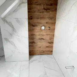 Wykończona łazienka z białymi płytkami imitującymi marmur na ścianach i podłodze, oraz drewnopodobnymi płytkami na jednej ze ścian, widoczny odpływ liniowy i przygotowane miejsce na baterię.