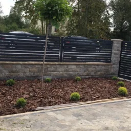 Nowoczesne ogrodzenie z poziomych, czarnych paneli na murku z szarej kostki, świeżo obsadzona rabata z korą i kulistymi krzewami, młode drzewko i wybrukowany podjazd.