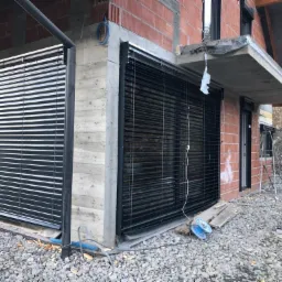 Elewacja surowego budynku z zamontowanymi zewnętrznymi żaluzjami fasadowymi w kolorze antracytowym, widoczne przewody elektryczne i drabina oparta o ścianę.