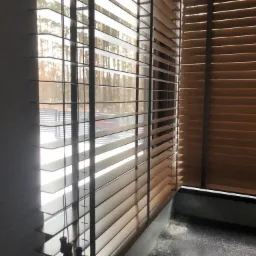 Brązowe poziome osłony okienne z drewna z widokiem na zewnątrz, częściowo zasłonięte, z widoczną strukturą drewna i sznurkami regulacyjnymi.