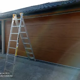 Montaż segmentowej bramy garażowej w kolorze drewna, z widoczną aluminiową drabiną opartą o bramę, na zewnątrz garażu z szarą kostką brukową.