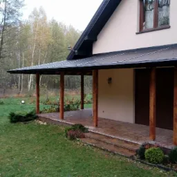 Zadaszony taras przy domu z drewnianymi słupami i kostką brukową, widok na trawnik i drzewa w tle.