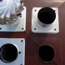 Widok z góry na cztery otwory w brązowym dachu: jeden z obrotową turbiną wiatrową, drugi z metalową rurą, trzeci z klapą wentylacyjną, czwarty pusty, z fragmentem rynny w tle.