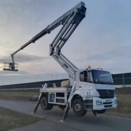 Biały samochód z wysięgnikiem koszowym marki Deko-Lift na rozstawionych podporach, stojący na asfalcie, z koszem uniesionym w górę na tle pochmurnego nieba i ekranów akustycznych.
