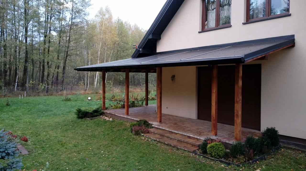 Zadaszony taras przy domu z drewnianymi słupami i kostką brukową, widok na trawnik i drzewa w tle.