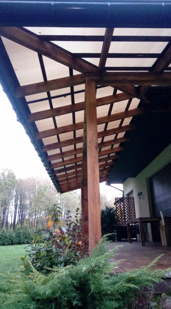 Drewniana pergola z przezroczystym zadaszeniem przy budynku, otoczona zielenią i krzewami ozdobnymi.