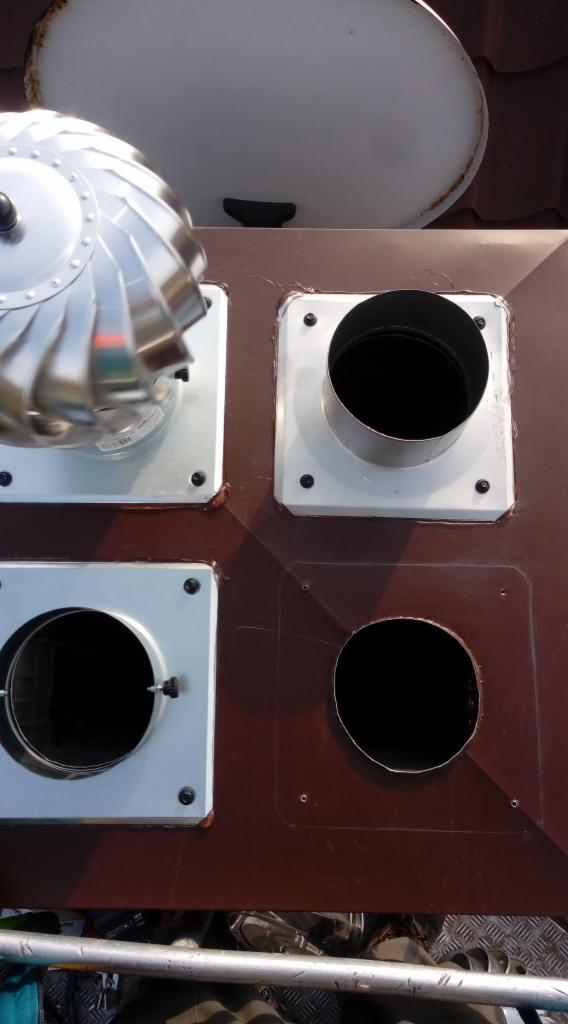 Widok z góry na cztery otwory w brązowym dachu: jeden z obrotową turbiną wiatrową, drugi z metalową rurą, trzeci z klapą wentylacyjną, czwarty pusty, z fragmentem rynny w tle.