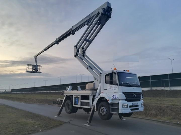 Biały samochód z wysięgnikiem koszowym marki Deko-Lift na rozstawionych podporach, stojący na asfalcie, z koszem uniesionym w górę na tle pochmurnego nieba i ekranów akustycznych.