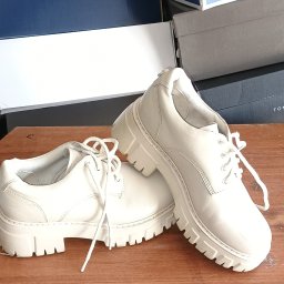 Hubert K. - Para białych, skórzanych butów typu derby na grubej podeszwie, ustawionych na drewnianym blacie na tle pudełek po butach różnych marek.