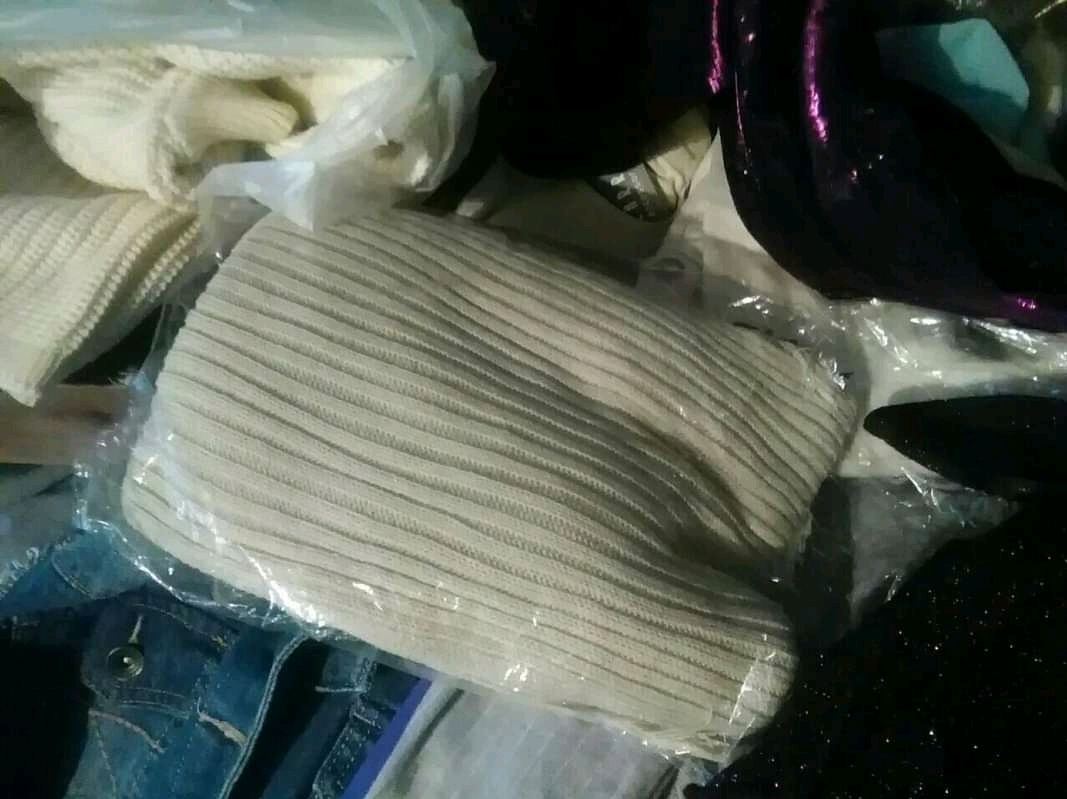 Stos ubrań, w tym kremowy sweter w prążki zapakowany w folię, dżinsowa kurtka z guzikiem i czarny błyszczący materiał.