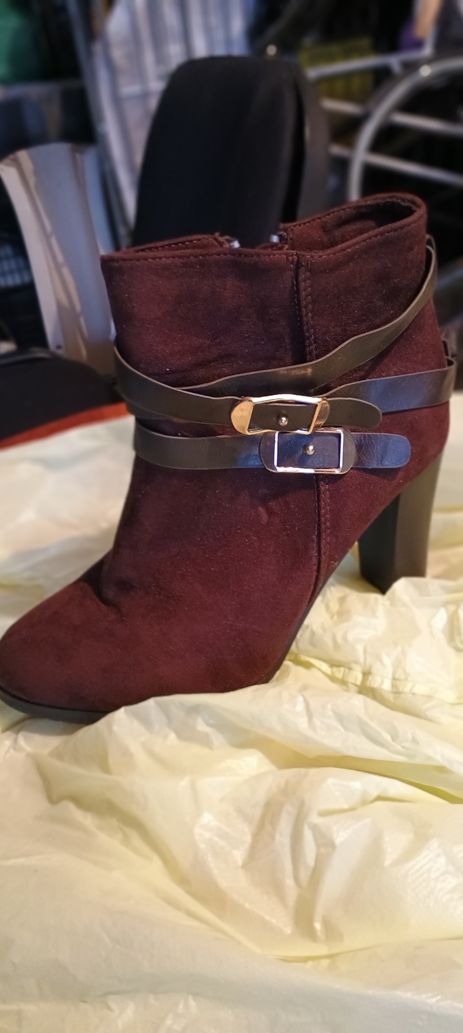 Burgundowy zamszowy botek na obcasie, ozdobiony dwoma paskami z klamrami, leżący na pogniecionym, jasnym podłożu.