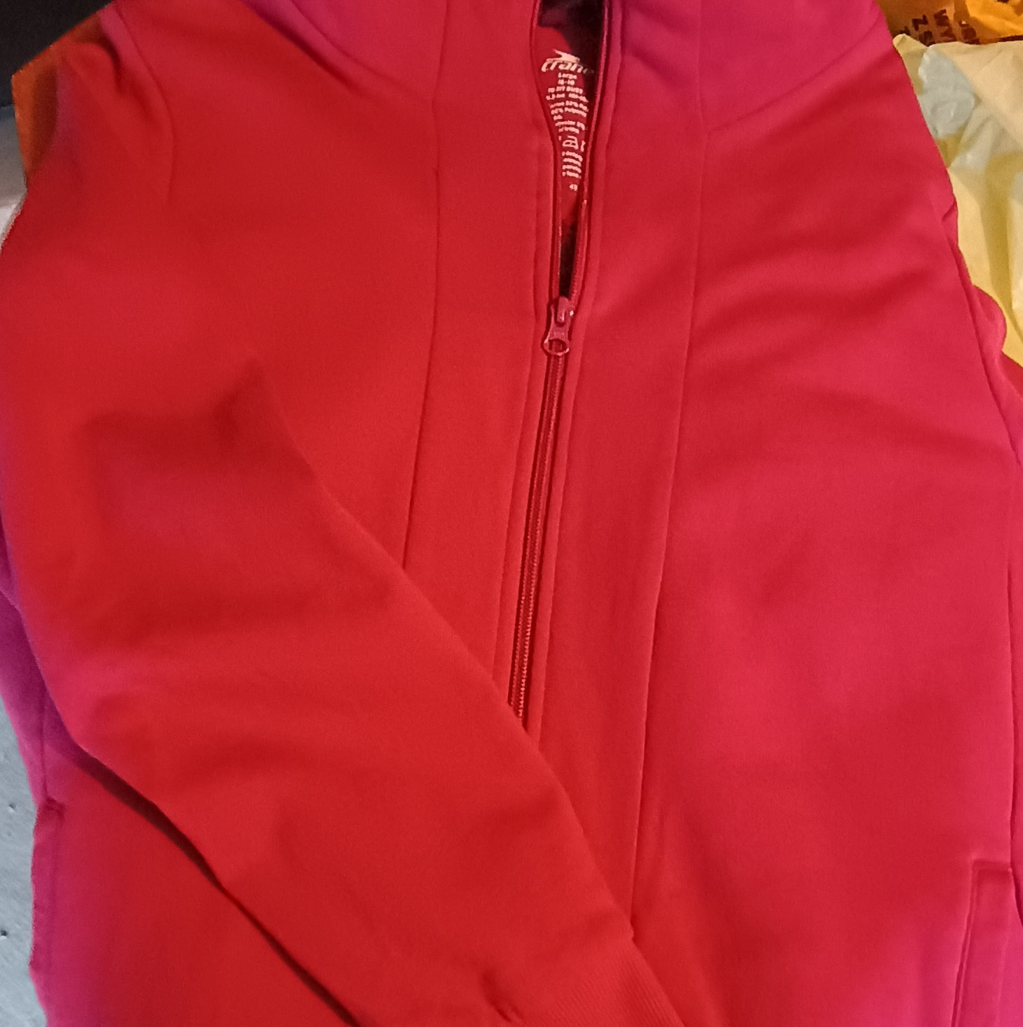 Czerwona rozpinana bluza sportowa z widocznym zamkiem błyskawicznym i metką producenta. Bluza leży na płaskiej powierzchni.