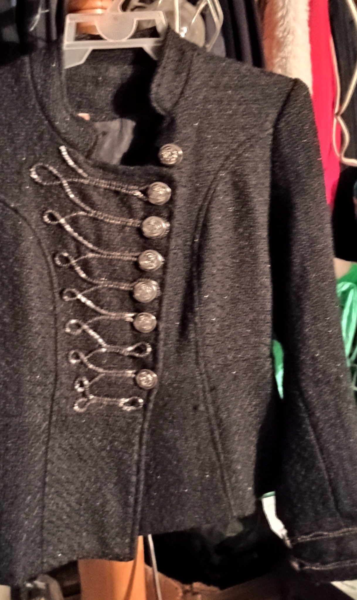 Czarna, krótka kurtka z ozdobnymi srebrnymi sznurkami i guzikami, wisząca na wieszaku w szafie.