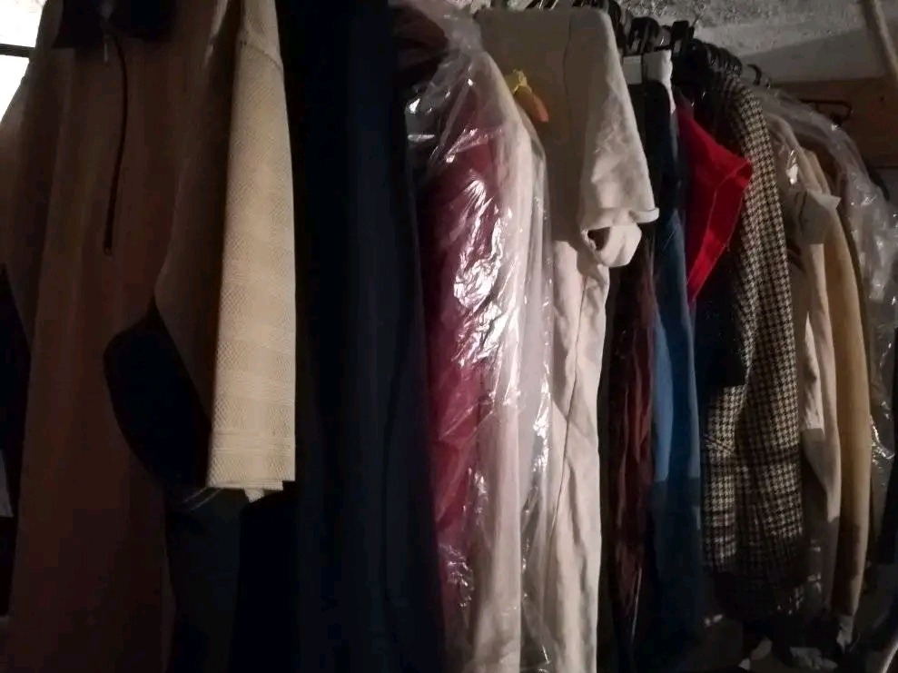 Wieszak z używanymi ubraniami, widoczne płaszcze, koszule i marynarki w różnych kolorach i wzorach, niektóre zapakowane w foliowe pokrowce.