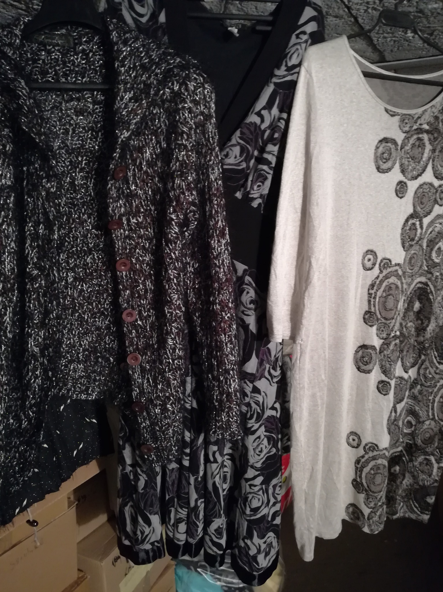 Ubrania damskie na wieszakach: dziergany kardigan z brązowymi guzikami, czarna bluzka i biała tunika z szarym wzorem w koła, wiszące obok siebie.