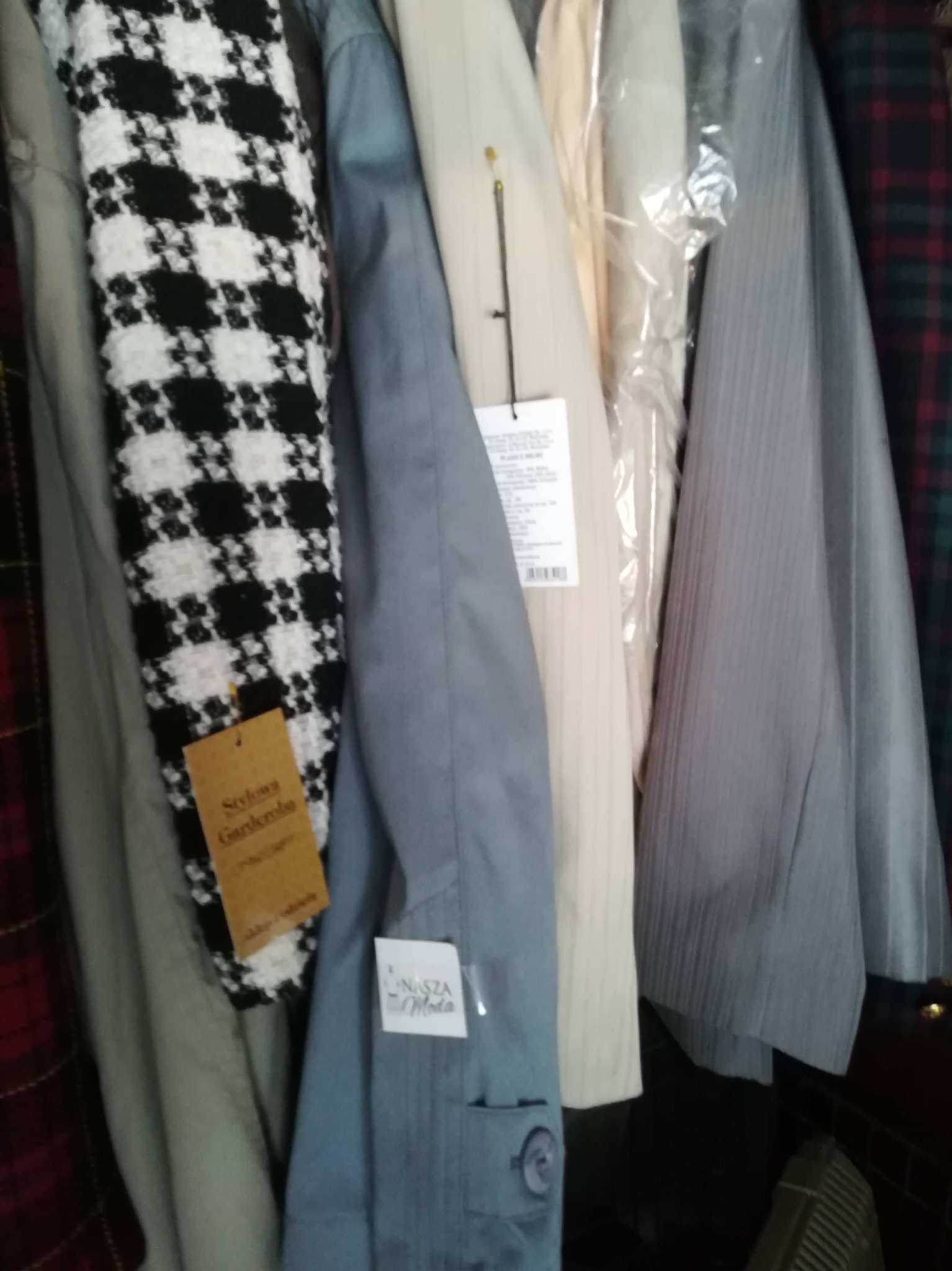 Ubrania wiszące na wieszakach, w tym płaszcz w pepitkę z brązową metką 'Stylowa Garderoba' i jasnoniebieska koszula z logo 'Nasza Moda'.