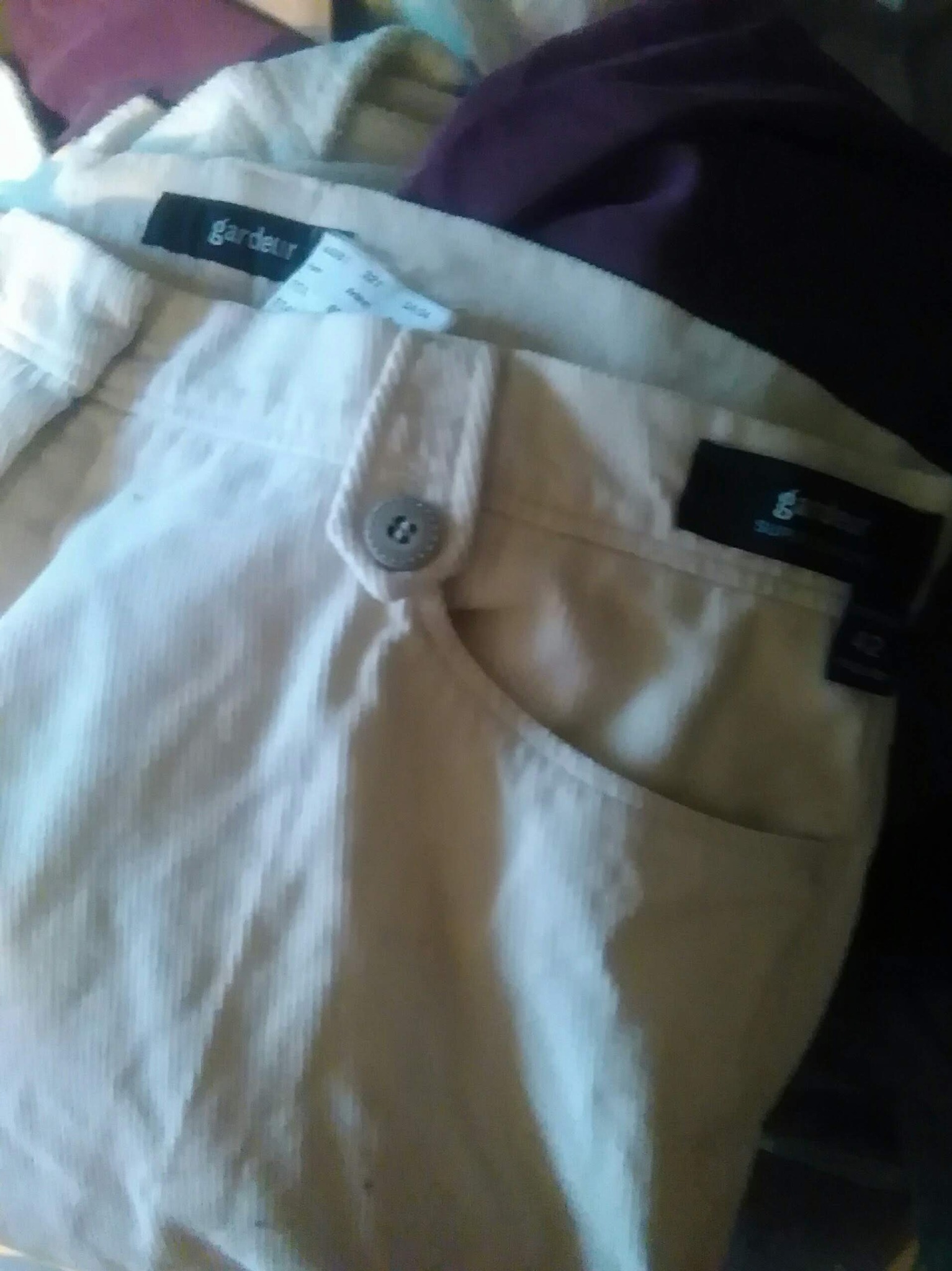 Beżowe spodnie marki Gardeur z widoczną kieszenią, guzikiem i metką, leżące na stercie innych ubrań.