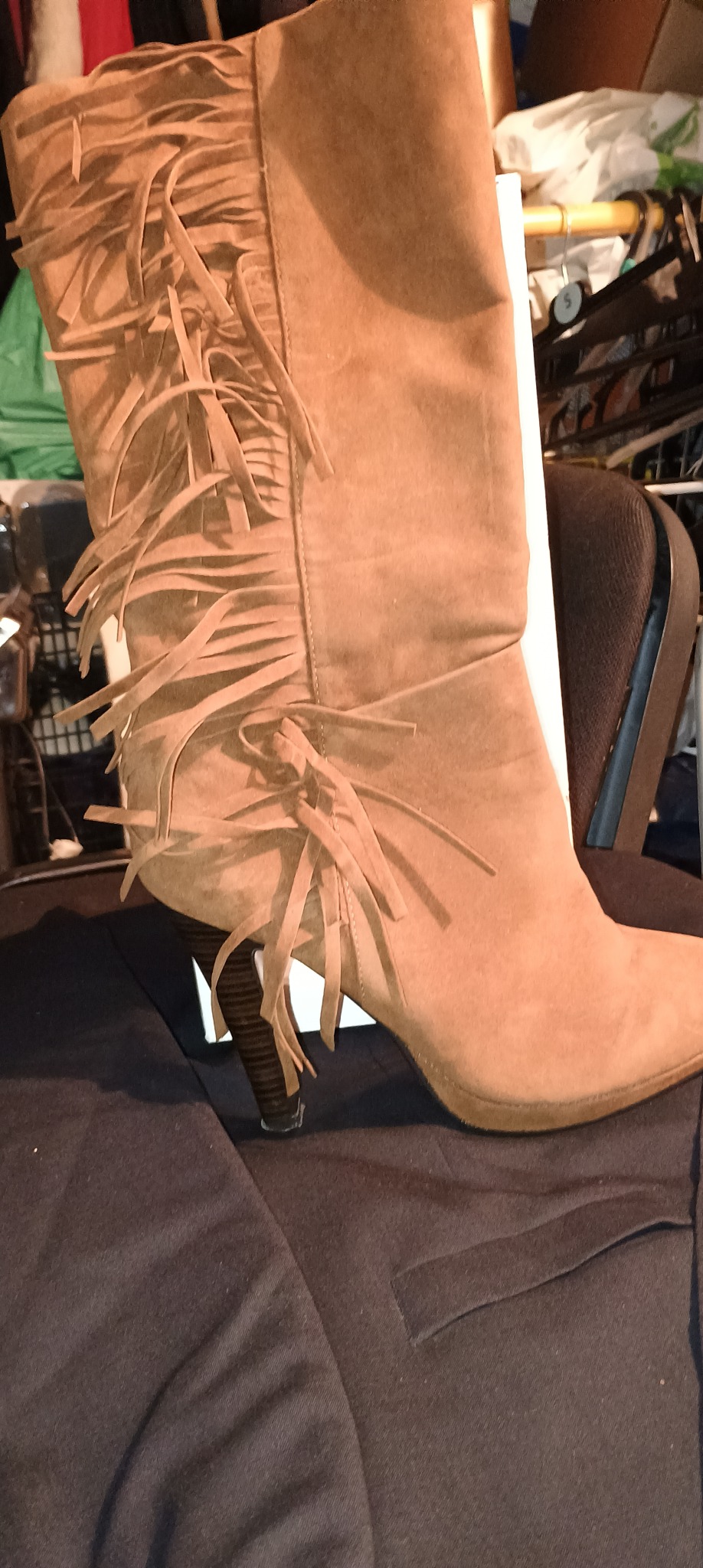 Brązowy zamszowy kozak z frędzlami na obcasie, ustawiony na ciemnym materiale, z widocznym chaotycznym tłem.