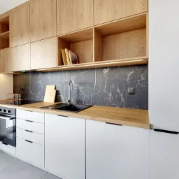 Nowoczesna kuchnia z białymi szafkami, drewnianym blatem i czarnym marmurem na ścianie, oświetlenie LED pod szafkami wiszącymi, płyta indukcyjna i piekarnik w zabudowie.