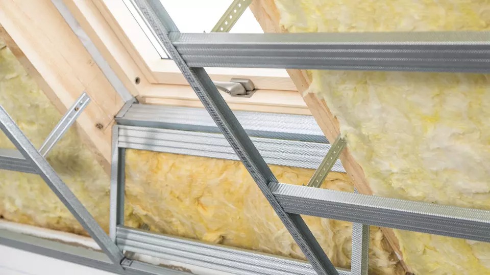 Ocieplenie poddasza wełną mineralną widoczne przez metalowy stelaż pod płyty karton-gips, okno dachowe w tle.